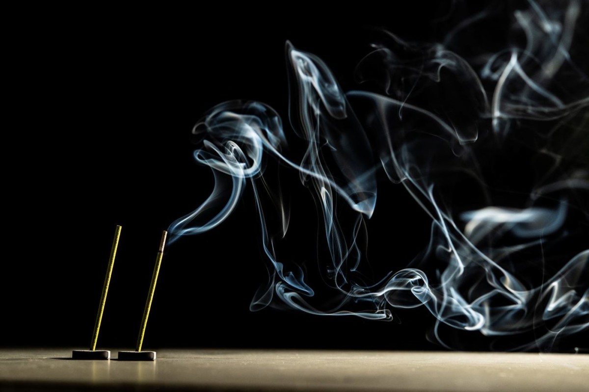 Image of two incense sticks on a table. The other one is burning and smoke rises from it.