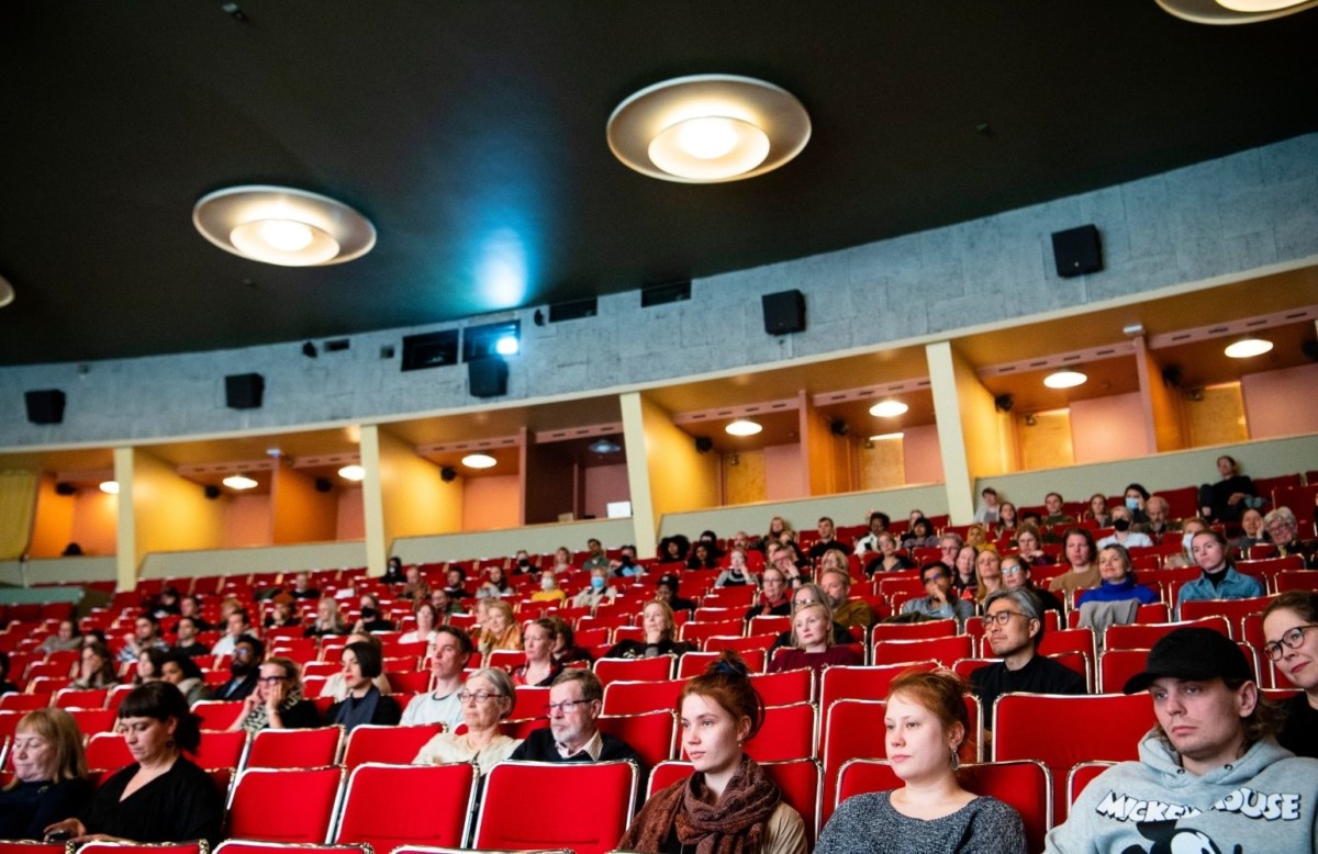 A cinema hall with red chairs and people sitting around the hall watching a film.