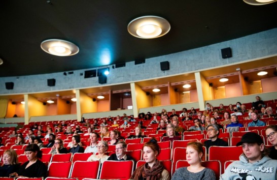 A cinema hall with red chairs and people sitting around the hall watching a film.
