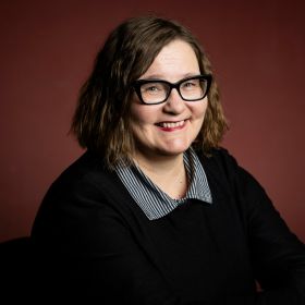 Image of a brown-haired smiling woman with glasses. She is wearing black shirt with striped collars. Backround is deep red.