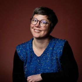 Image of a smiling woman with glasses. She has dark brown short hair and she is wearing a dark blue vest with black shirt. Background is deep red.