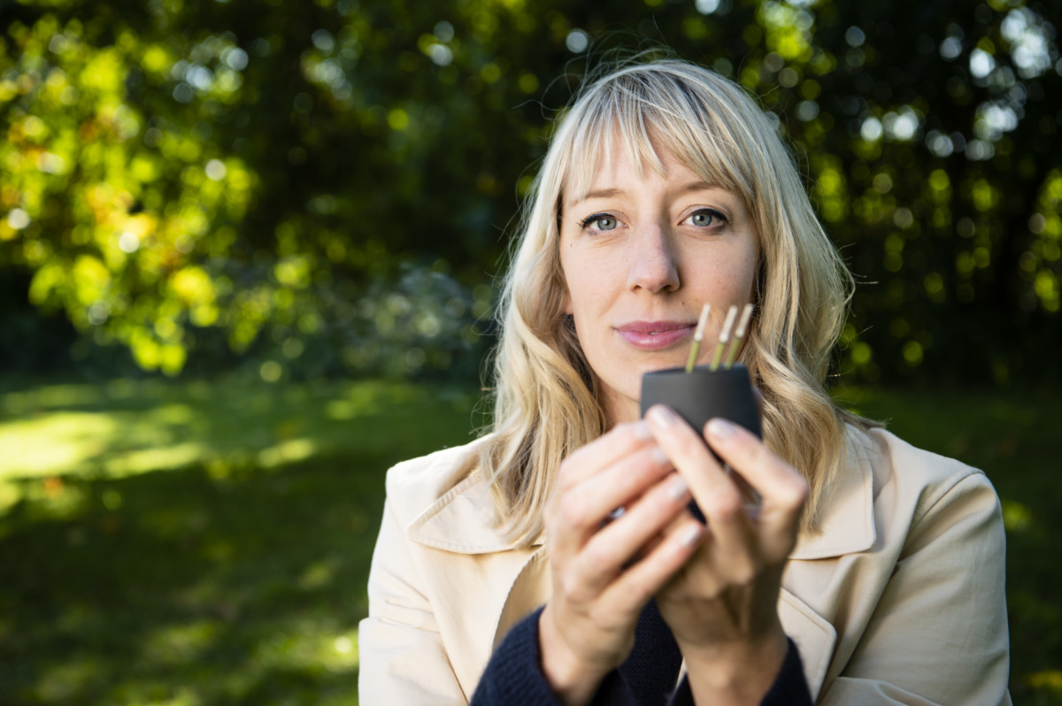 Image of Katie Paterson, a woman with blond mid-length hair standing outside in a park, smiling and holding a cup of three incense sticks in her hands, facing towards the camera.