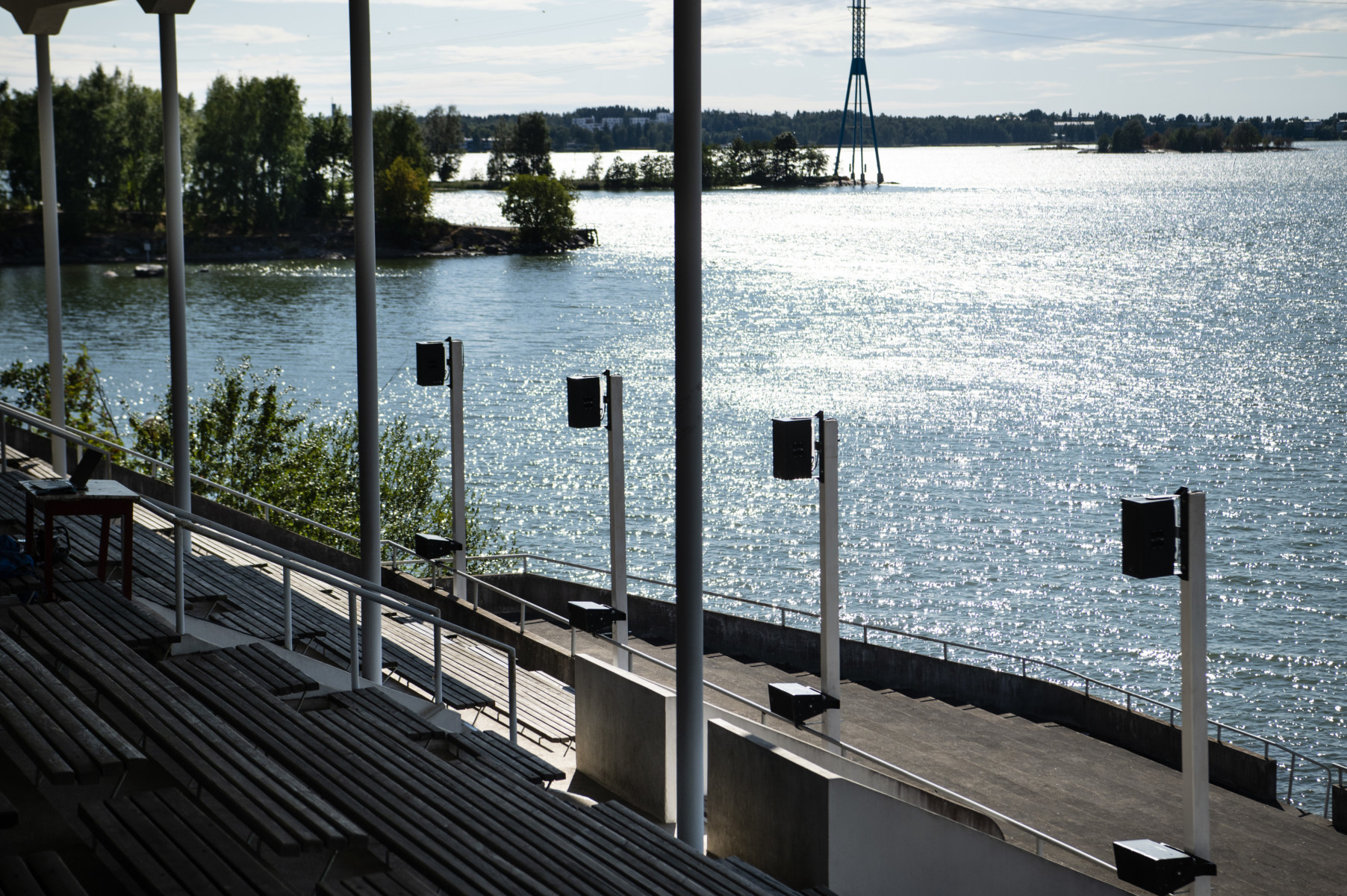 Image of a rowing stadium in Helsinki in a sunny day. Four big loud speakers are facing the stadium and behind them is a sea view glimmering in sun.