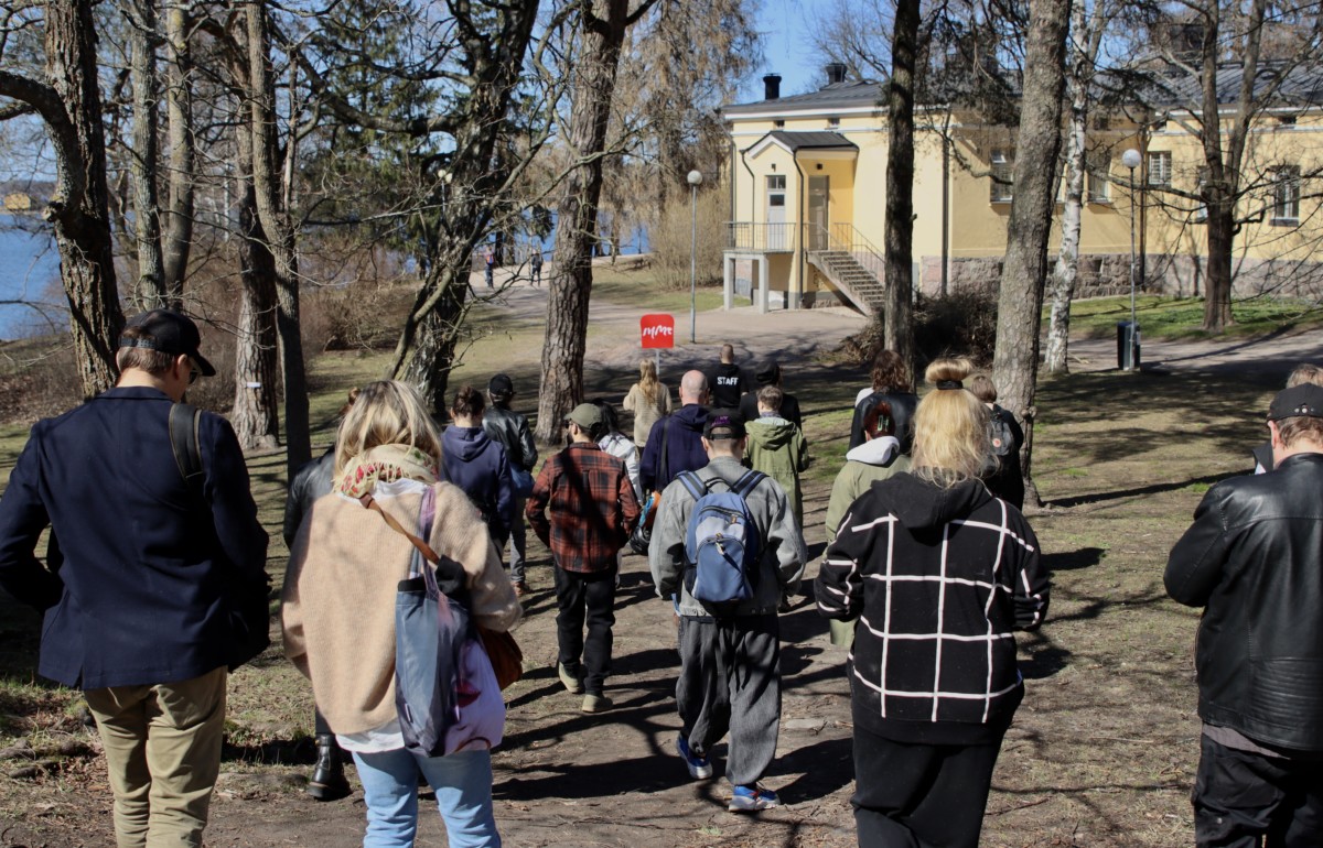 A group of people walking outside in a park of an old yellow building on a sunny day. One person is holding a red sign reading IHME in white letters.