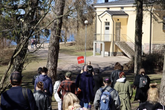A group of people walkin outside in a park of a yellow old building on a sunny day. One person is holding a red sign that reads a text IHME in white letters.