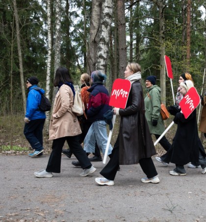 A group of people walking in a forest on a cloudy day. Three persons are holding red signs that read text IHME in white letters.