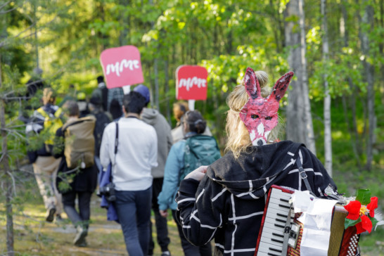 A group of people walking in the forest, backs towards the camera. Two people are carrying red signs that read IHME in with letters.