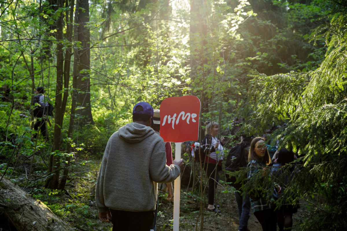 Photo: A person is walking in a green forest with an IHME sign in their hand. There is a ray of light coming down through the green leaves, and there is hopefulness in the photo. Maybe environmental handprint is being created right at this moment?