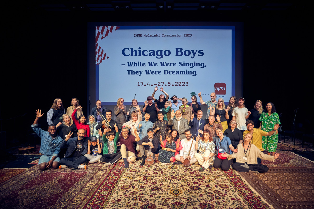 Kuvassa on Chicago Boys -teoksen osallistujia teoksen viimeisissä julkisissa harjoituksissa kulttuurikeskus Stoan teatterisalissa. Iso ryhmä hymyileviä ja nauravia ihmisiä kädet ylhäällä. Lattialla on värikkäitä persialaisia mattoja ja taustalla valkokankaalle heijastettu teksti IHME Helsinki 2023 -teos, Chicago Boys - While We Were Singing, They Were Dreaming 17.4. - 27.52023.