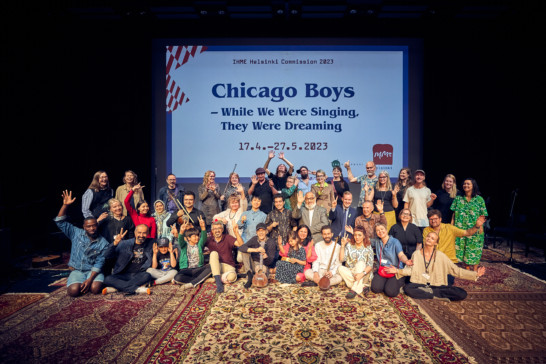 Image of a group of people smiling, laughing and waving hands high together at the final public rehearsal of Chicago Boys - While We Were Singing, They Were Dreaming in the theatre hall of Stoa Cultural Centre. The group is standing and sitting on colorful persian carpets. Behind them is a screened text saying IHME Helsinki Commission 2023 Chicago Boys - While We Were Singing, They Were Dreaming 17.4. - 27.5.2023.