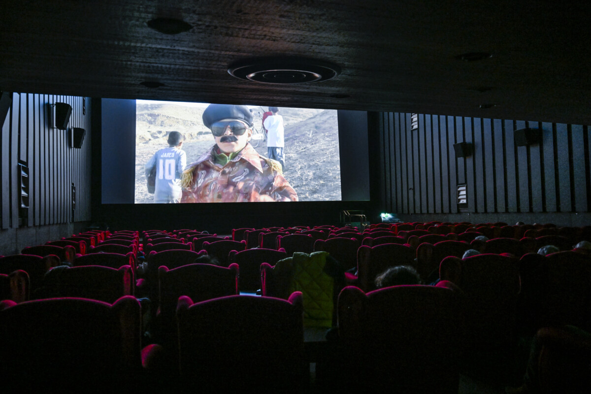Kuvassa on elokuvasali, jossa on punaisia nojatuoleja ja valkokankaalla pyörii elokuva, jossa on lapsia aavikolla.