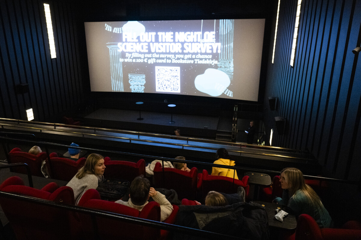 Image of a cinema hall viewed from the balcony, People sitting in red armchairs and waiting for the film to begin.