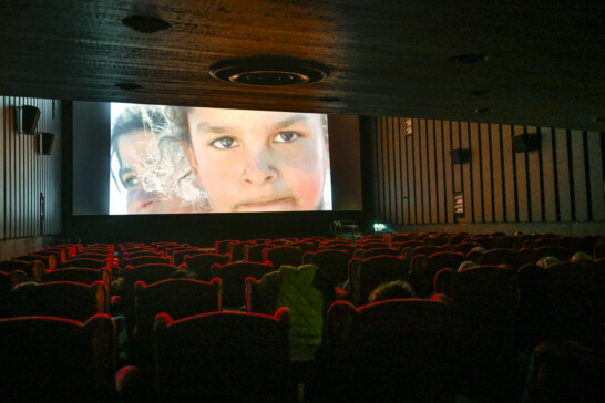 Kuvassa on elokuvateatterin sali, jossa on punaisia nojatuoleja ja valkokankaalla lapsen kasvot.