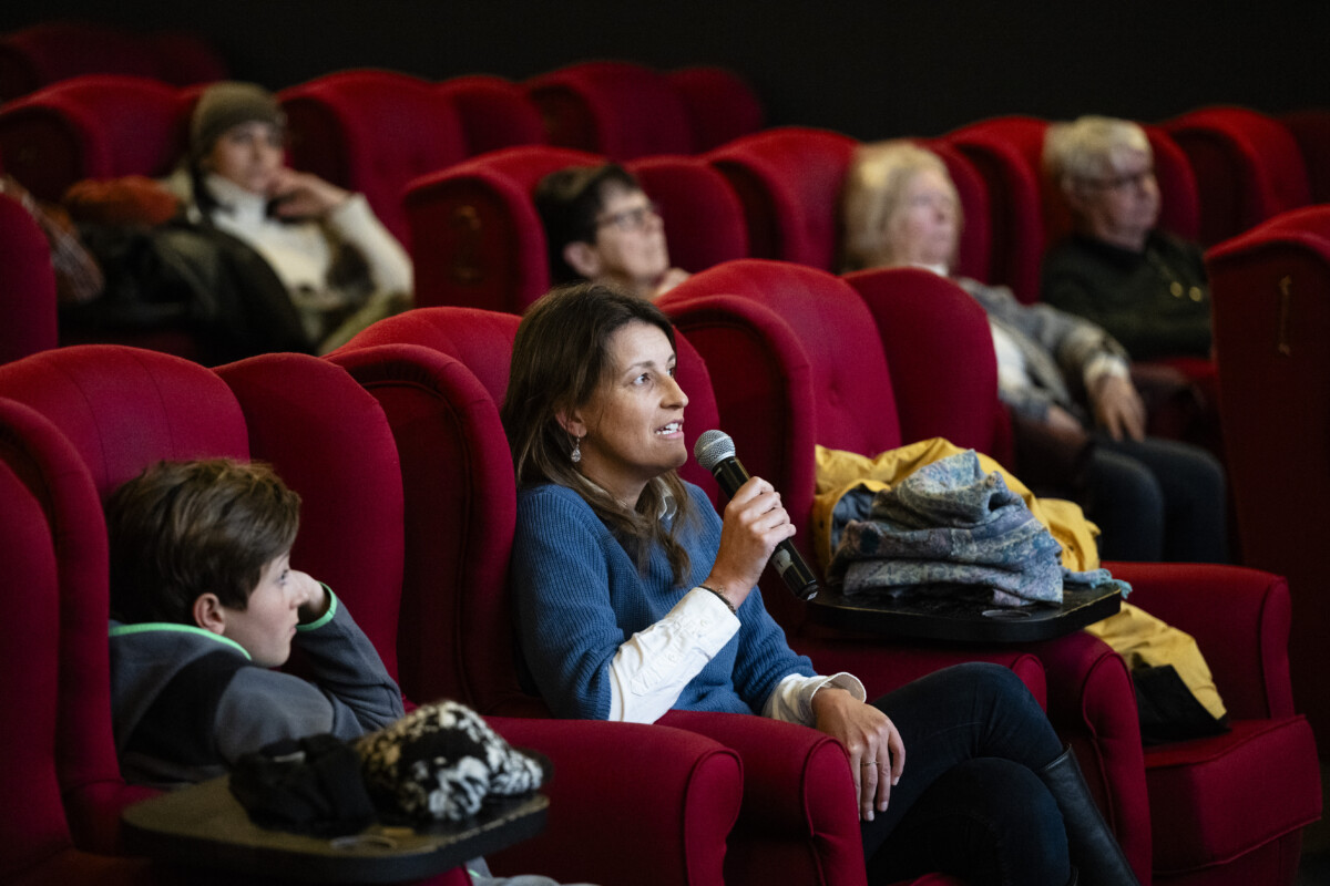 Kuvassa on elokuvateatterin sali, jossa on punaisia nojatuoleja ja ihmisiä istuu kuuntelemassa keskustelua. Yksi henkilö kysyy kysymystä mikrofoni kädessä.