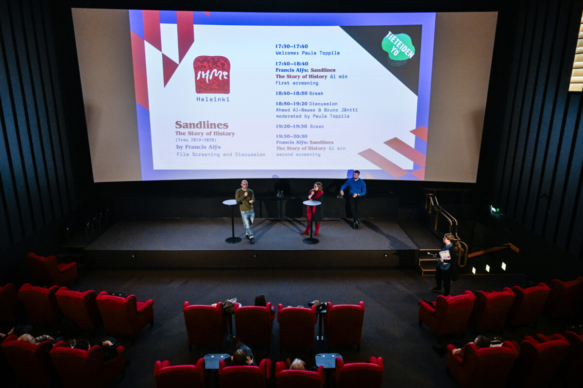 Image of a cinema hall viewed from the balcony, people sitting in red armchairs and three persons standing on the stage and talking.