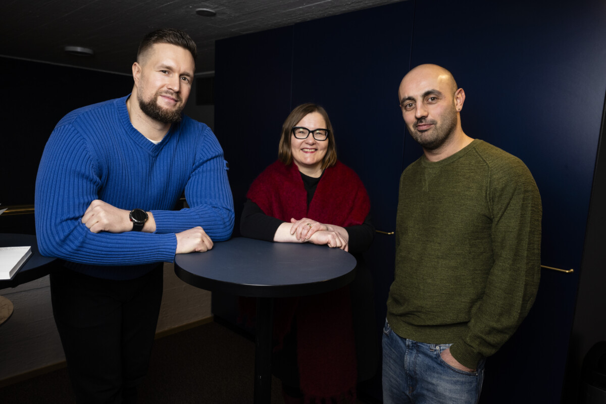 Image of a three smiling people standing around a round bar table. Bruno Jäntti has short brown hair and beard and he is wearing a blue sweater and black trousers. Paula Toppila has glasses and brown mid-length hair. She is wearing a black dress with a red scarf. Ahmed Al-Nawas has bald head and stubble. He is wearing green sweater and jeans.