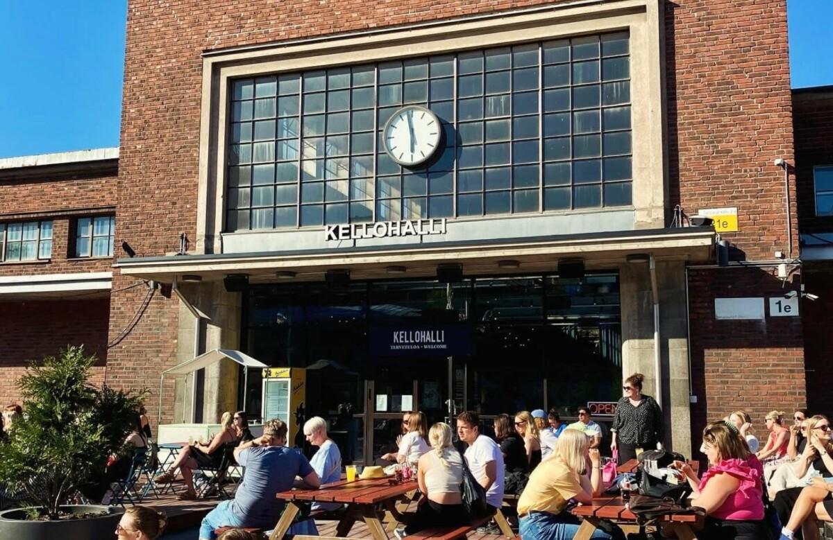 Image of a red brick building on a sunny day. The building has a large glass wall and a canopy on top of which reads Kellohalli. There are people sitting outside at the terrace of the building.