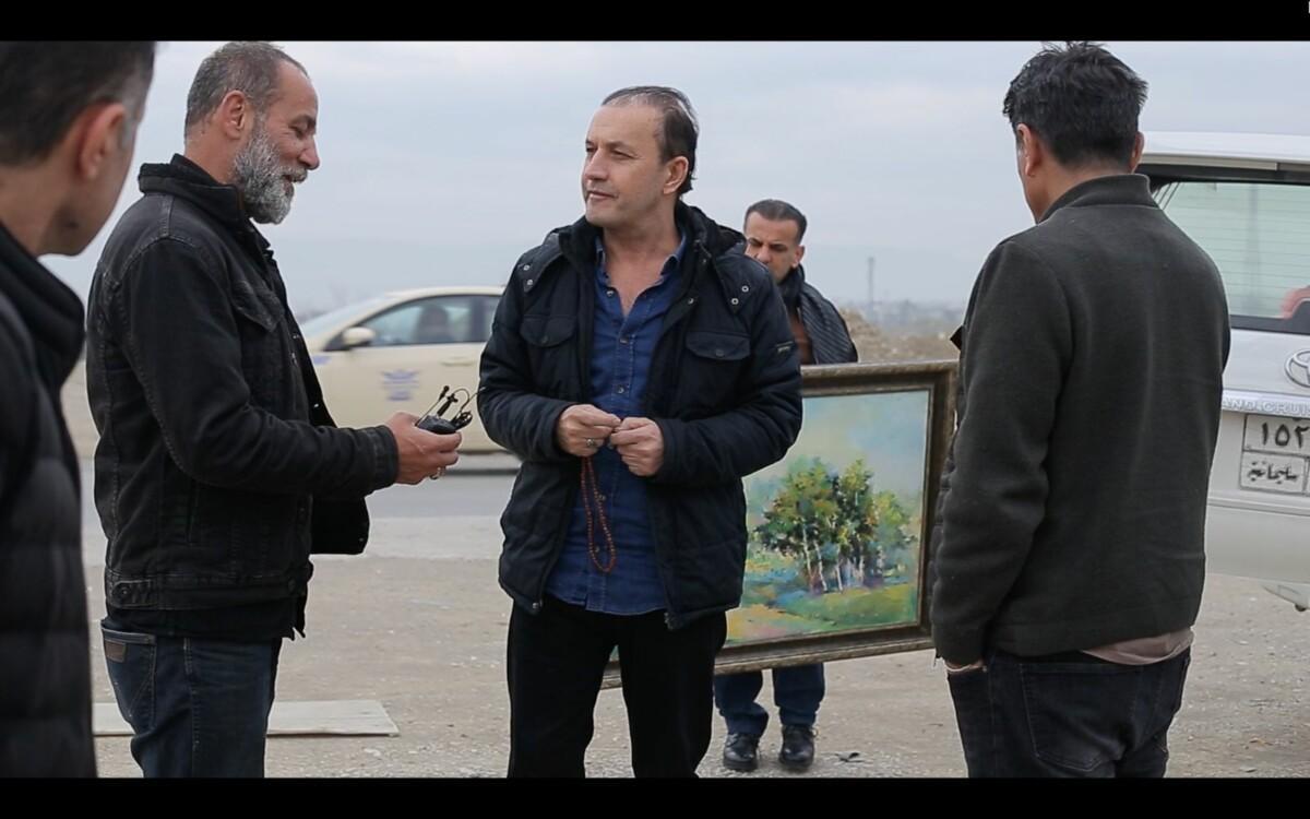 Four men standing outside in a parking lot and talking to each other. On man is holding a landscape painting with a view of a green forest.