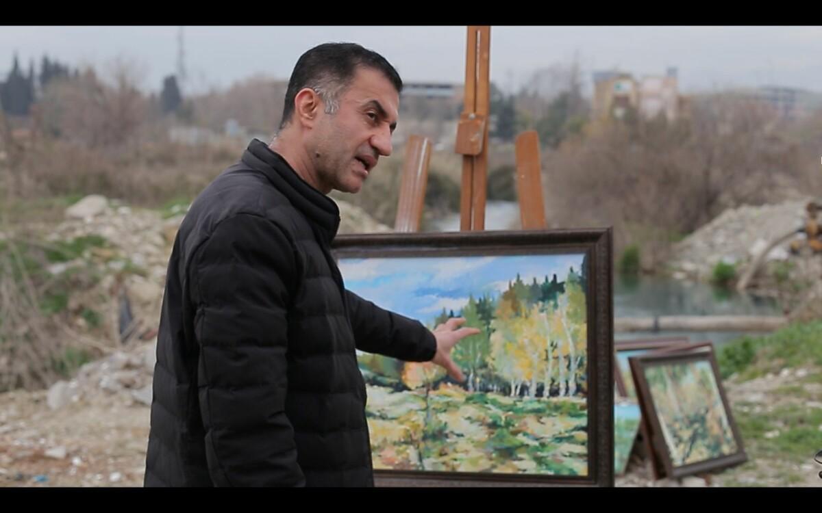 A dark haired man in a black coat standing outside in front of a landscape painting and pointing to it while talking. Behind a scenery of leafless trees and buildings. Cloudy day.