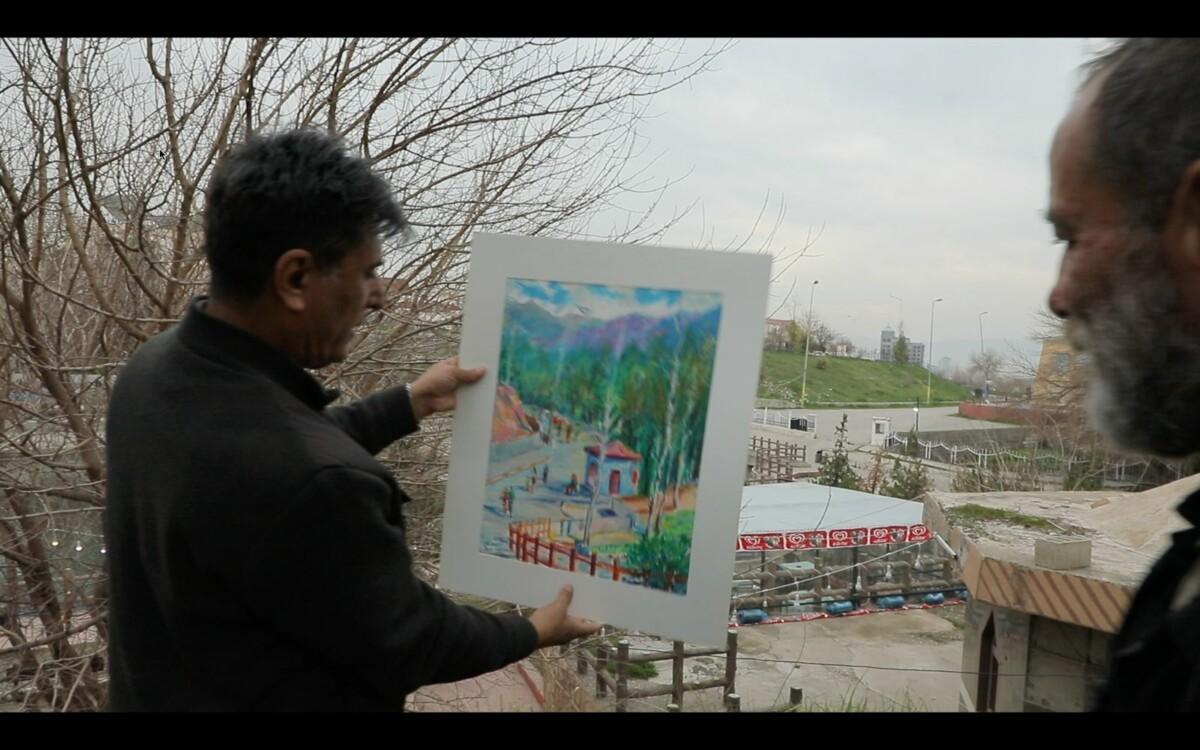 A dark haired man in a brown coat standing outside holding a painting in his hand in front of a scenery. The painting is a landscape painting with green forests. Another man with beard is standing a side and looking at the paiting.