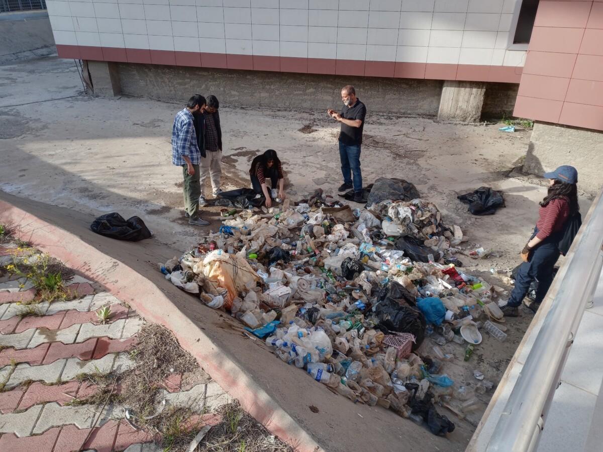 A group of people standing outside on a sunny day at a street between buildings. The people are surrounding a pile of plastic bottles and other trash.
