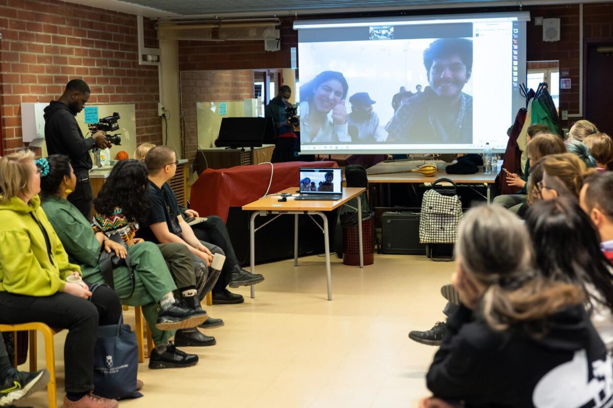 A group of people in Helsinki sitting in a class room and watching a wide screen with an image of another group of people in Sulaymaniyah.