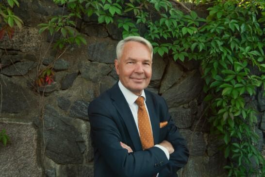 Image of Pekka Haavisto, a grey haired man smiling towards the camera with hands crossed. The man is wearing a black suite with an orange tie. He stands in front of a rock wall that is filled with bine.