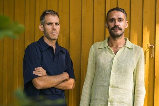 Cooking Sections' artists Alon Schwabe and Daniel Fernández Pascual, two men standing in front of a yellow wooden wall. Alon is wearing a blue shirt and standing hands crossed. Daniel in wearing a light yellow shirt. Both are looking straight to the camera.