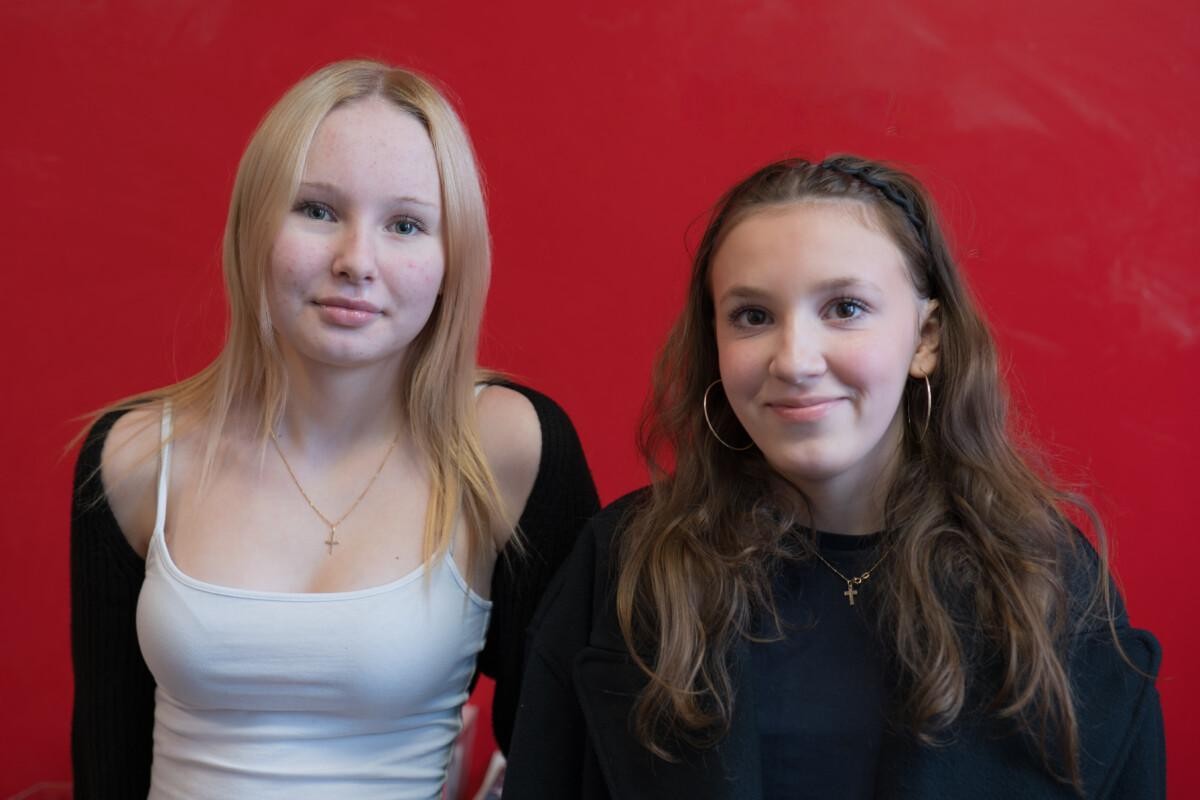 Two young girls standing and smiling in front of a red wall. The othe one has long blond hair and the other one long bronw curly hair.