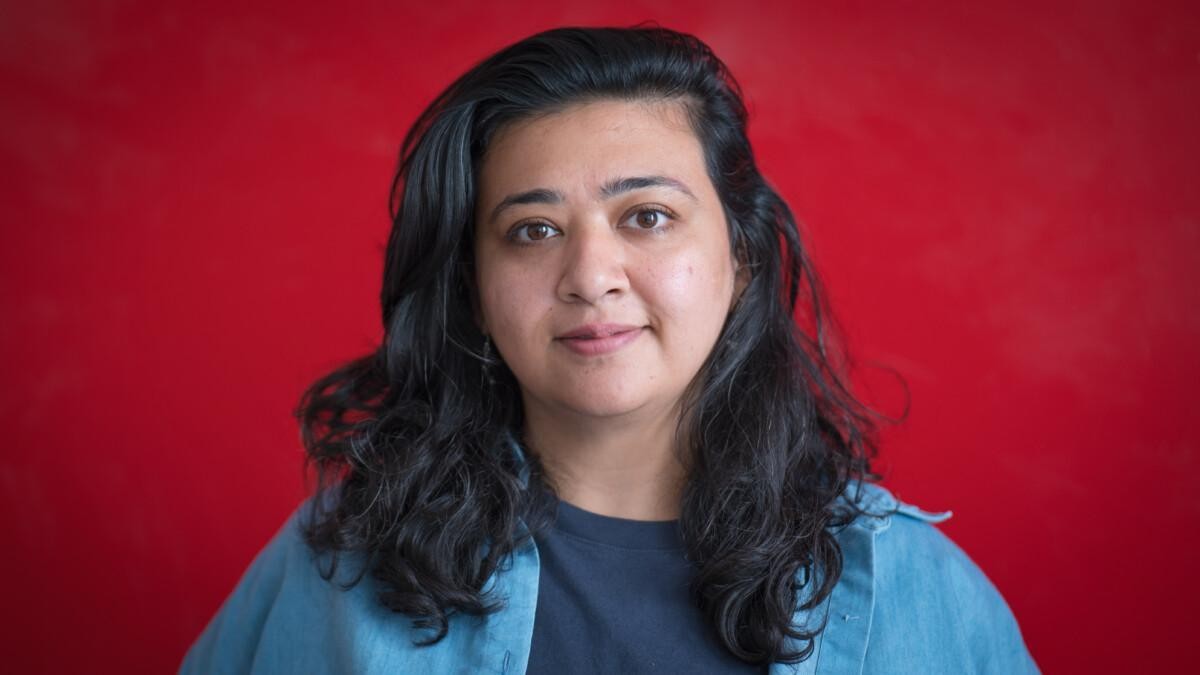 Image of Zara Asgher, a dark-haired, smiling woman. Standing in front of a red wall. She is wearing light blue collar shirt and dark blue t-shirt.