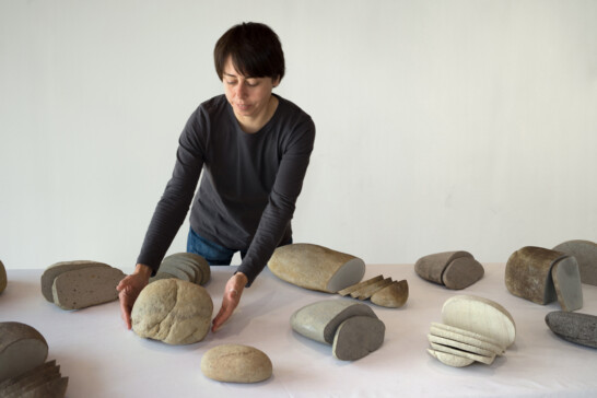 An image of a table with white table cloth. Covered with stones of different sizes resembling of breads. The artist Zhanna Kadyrova is placing the stones on the table. She has short dark hair, dark long sleeved shirt and jeans.