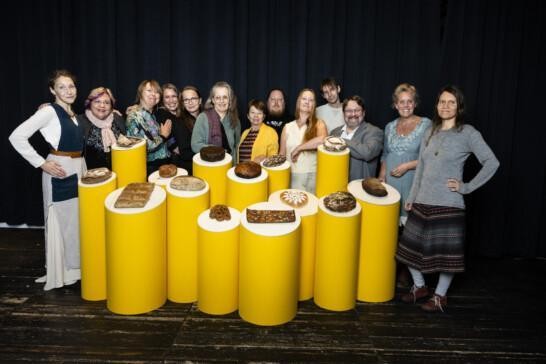 A group photo of fourteen people standing behind yellow stands holding different types of bread.