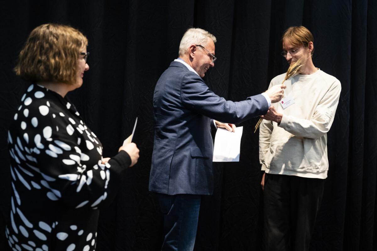 The picture shows two people shaking hands. One is wearing a blue-grey suit and has grey hair. The other is wearing a light sweatshirt and has brown hair.