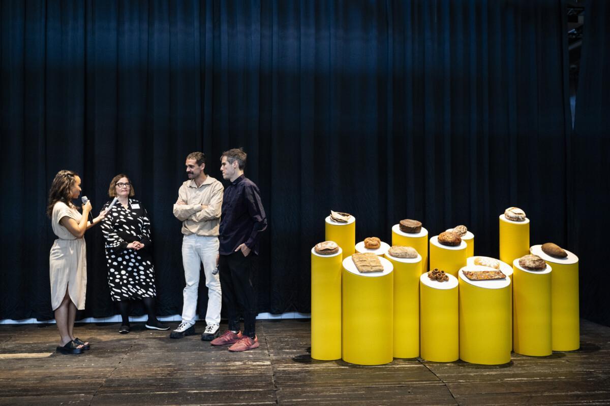 The picture shows four people on the left edge of the stage and on the right edge is a bread display of yellow stands and loaves of bread.