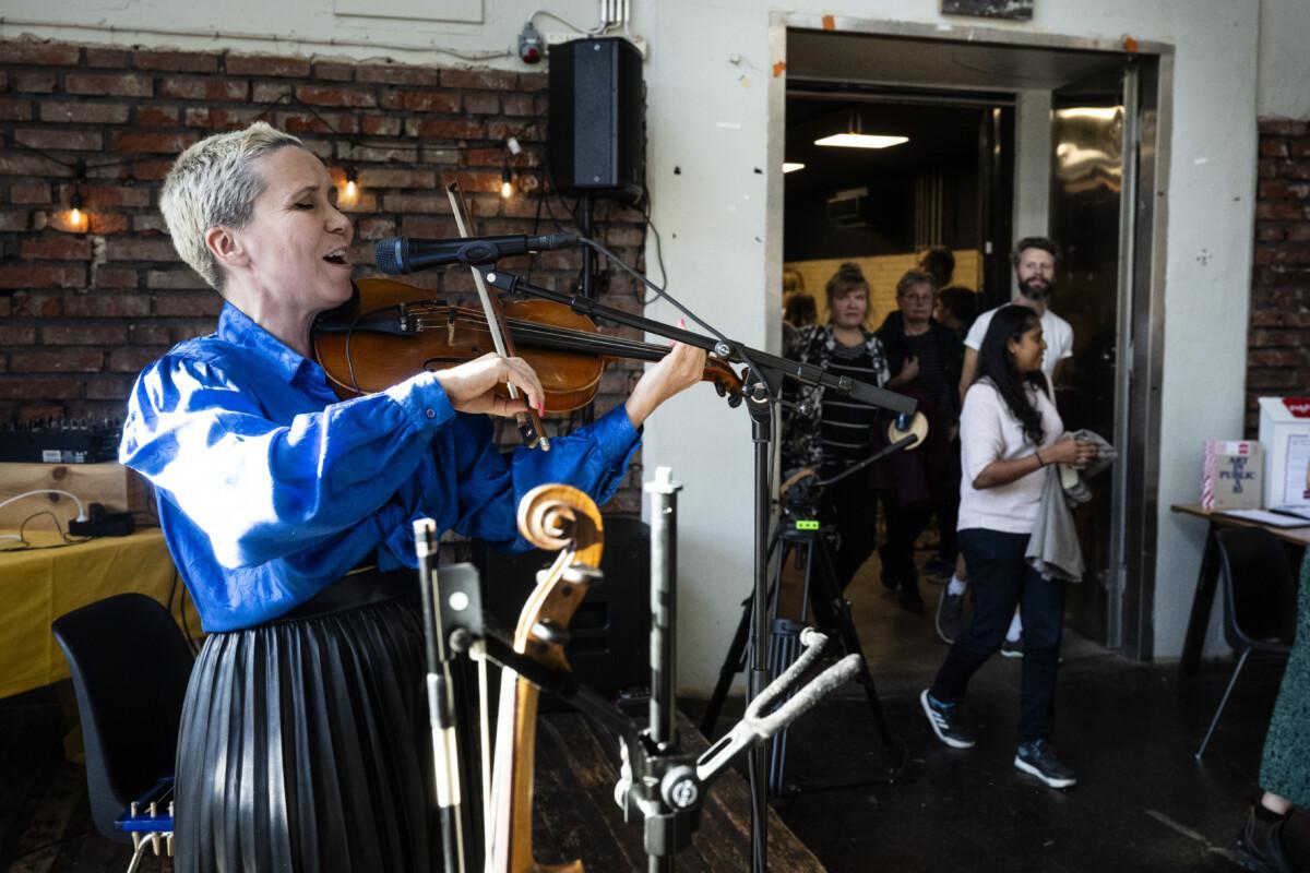 The picture shows a short-haired woman in a blue shirt and black skirt singing and playing the violin for the audience.