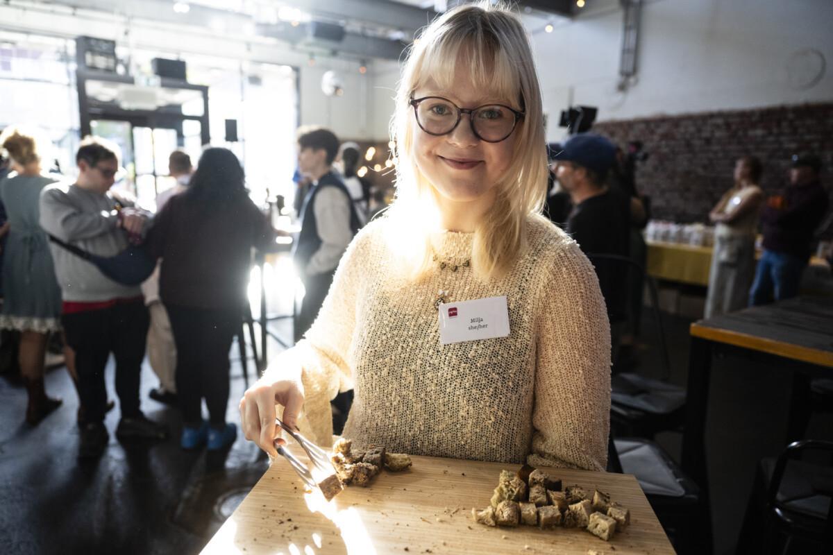 The picture shows a smiling young woman with blond hair and glasses, holding pieces of bread on a wooden tray to taste.