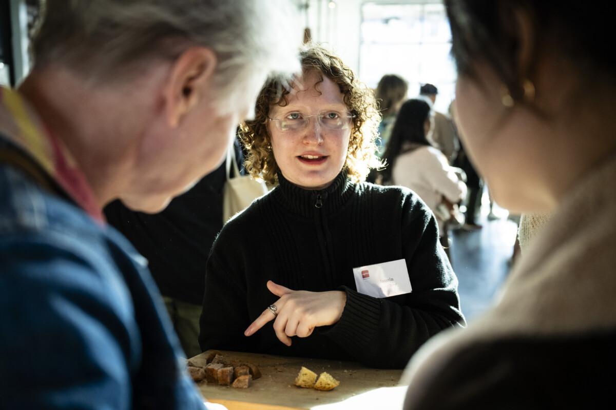 The picture shows a smiling, bespectacled, dark-haired person with curly hair serving a bread to two other people.