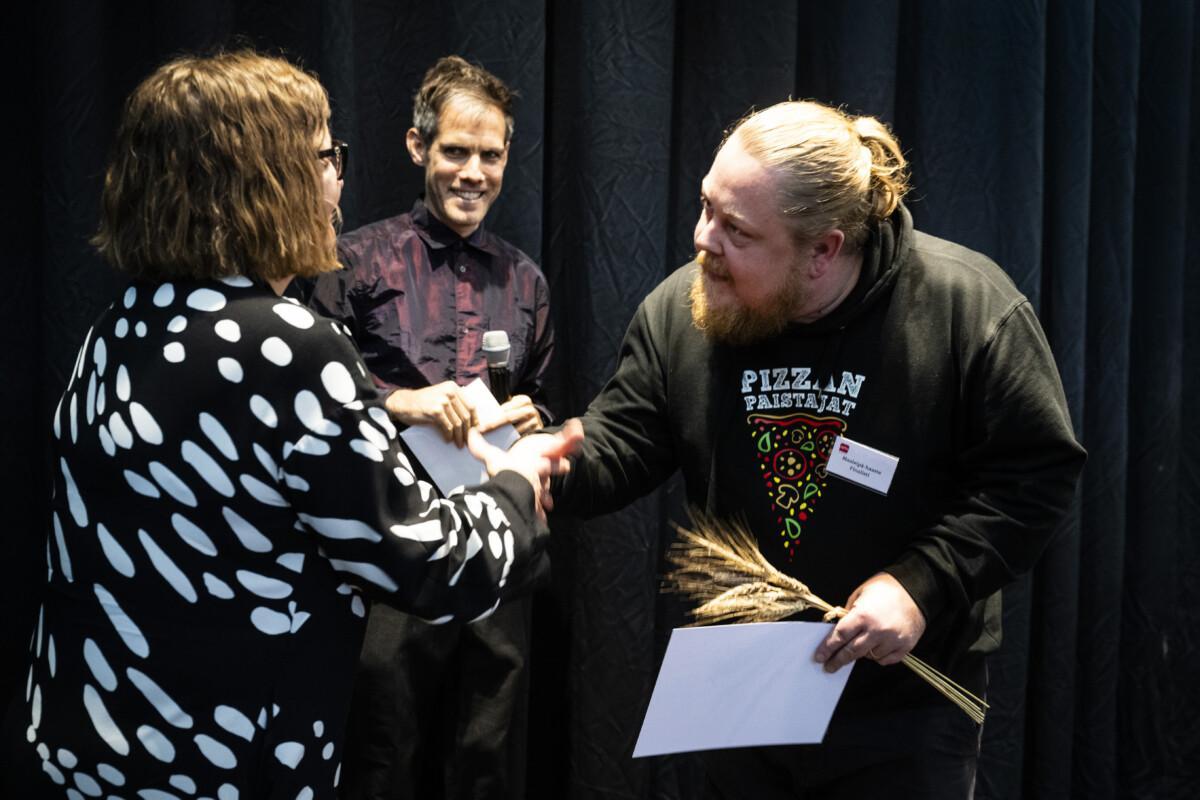 The picture shows three people, two shaking hands and a third smiling in the background.