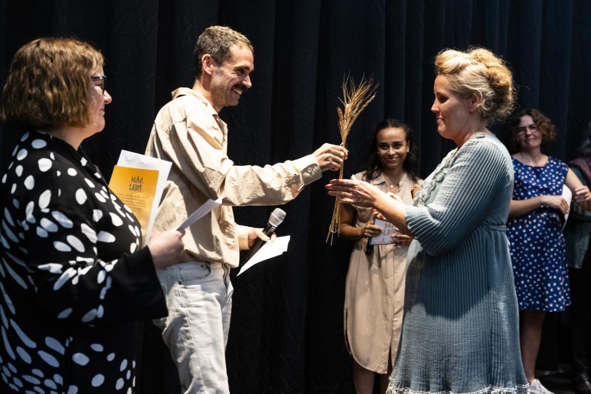 Three people are pictured, one of whom is handing Malin Furu, a blonde-haired woman in a blue dress, a congratulatory bouquet made of cereal corsages. A third person is smiling beside her.