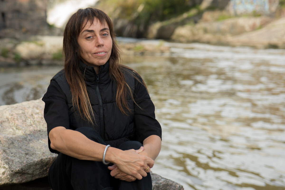 Image of Zhanna Kadyrova. A dark haired woman wearing black clothes and sitting on a rock by a river.