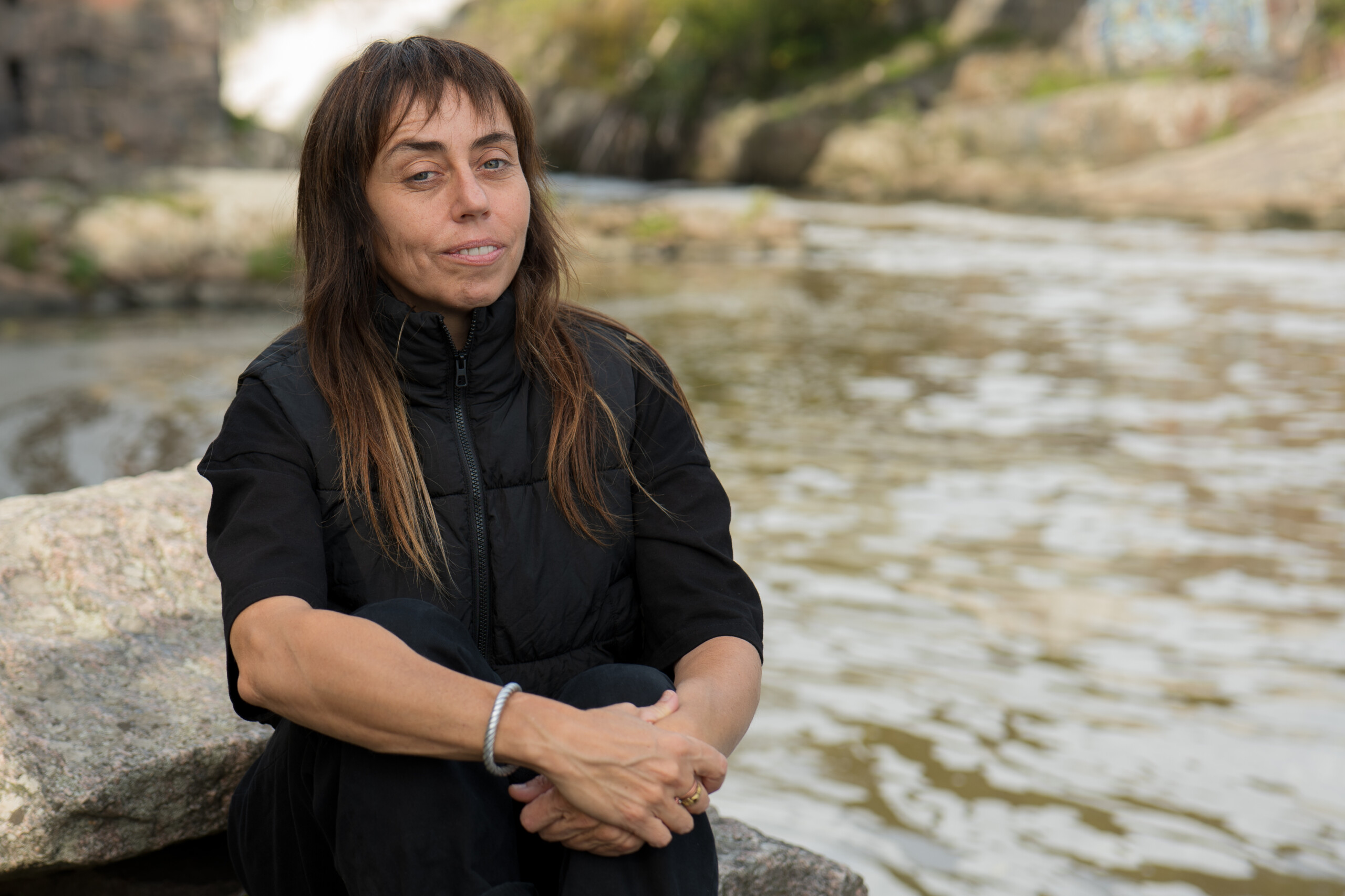 Image of Zhanna Kadyrova. A dark haired woman in black clothes sitting on a rock by a river.