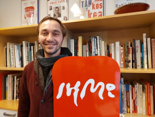 Image of Halil Kisyo, a young smiling man with dark brown hair and red sweater. He is standing in front of a bookshelf and holding a bright red sign that reads IHME in white big letters.