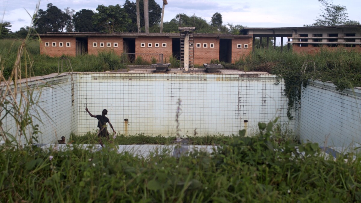 The picture shows old buildings among the sprawling vegetation and an empty swimming pool with a child playing on the edge.