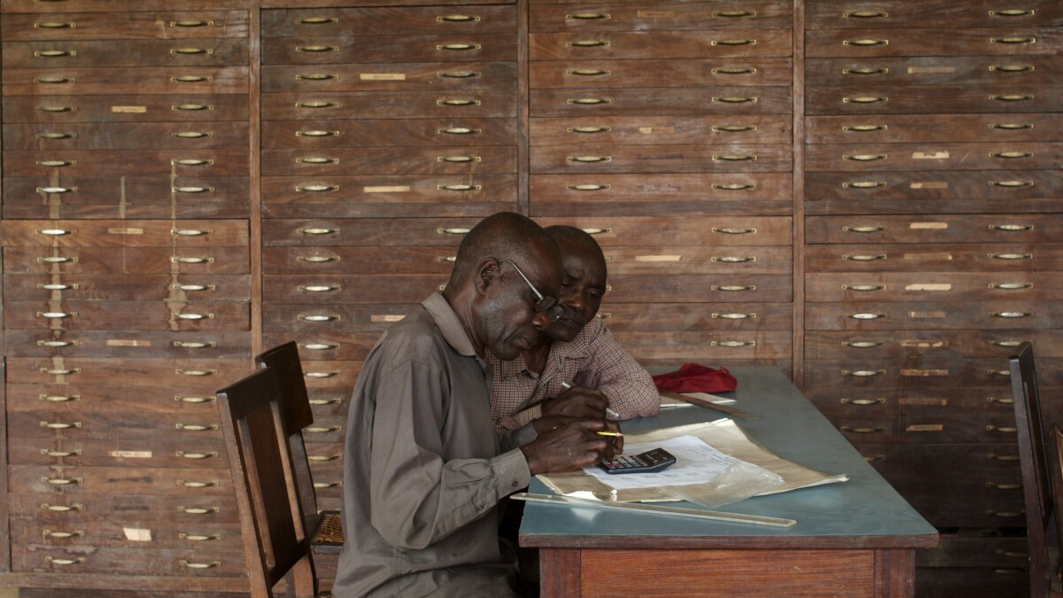 The picture shows two African men sitting at a table, working with old papers and a calculator. In the background is a filing cabinet.