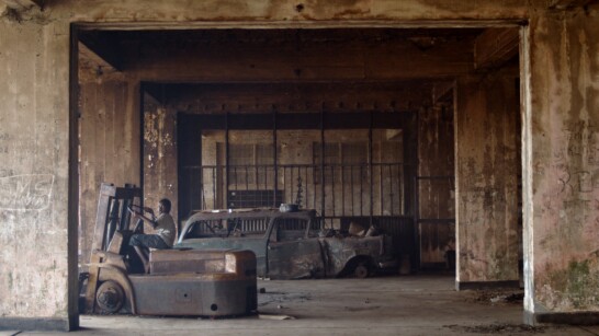 The picture shows a dilapidated industrial building with old, rusting agricultural machinery and cars. A dark-skinned child sits on one of the machines, looking at the camera.