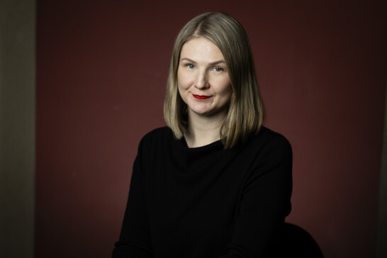 Pinja Valja, a media artist specializing in climate expertise, joins the IHME Helsinki team. Image of a person with shoulder-length blonde hair, red lips, and a black shirt. The background is a dark red wall.