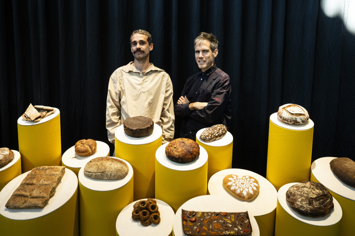 In the picture are Cooking Sections' artists Daniel Fernández Pascual and Alon Schwabe. Two men smiling and standing behind an installation that contains different sized pedestals with various breads on them.