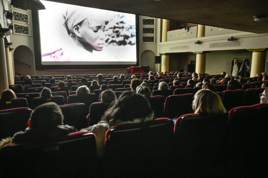 The image shows a movie theater auditorium with people seated. A film is playing on the big screen.