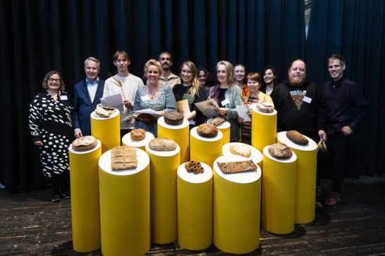 A group of smiling people standing behind an installation of different kinds of breads on different pedestals.