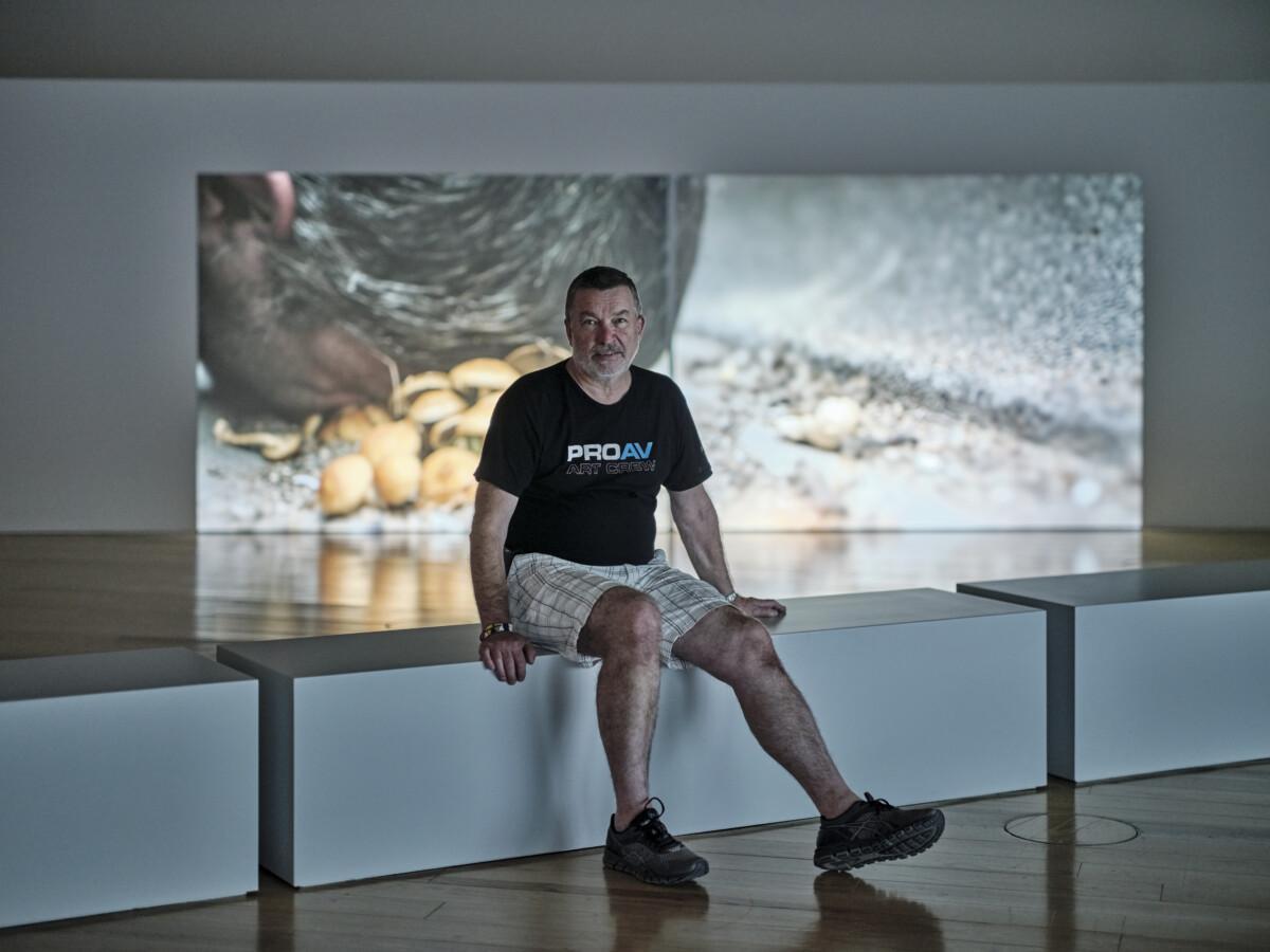 Kuvassa on Jorma Saarikko istumassa näyttelytilassa videoteoksen edessä. Hän on keski-ikäinen lyhythiuksinen mies, jolla on päällä musta t-paita, vaaleat shortsit ja mustat lenkkarit.